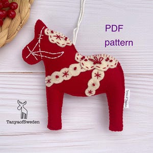 May include: A red felt Dala horse decoration with white embroidered details and a hanging string. The ornament features the text "PDF pattern" and the logo "TanyaofSweden".