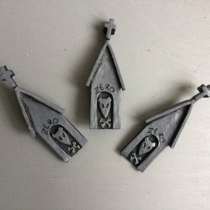 Puede incluir: Tres figuras grises en miniatura con forma de casa con una cruz en la parte superior. Cada una tiene la palabra "ZERO" y una cara de perro de dibujos animados con huesos cruzados. Las figuras están dispuestas sobre una superficie gris.
