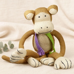 Könnte beinhalten: Ein gestricktes Affenspielzeug mit hellbraunem Körper sowie cremefarbenem Gesicht, Händen und Füßen. Der Affe hat einen einfachen gestickten schwarzen Mund und Augen und trägt einen zweifarbigen Schal mit einer violetten und einer lindgrünen Seite.