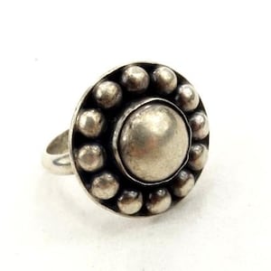 May include: A silver ring featuring a central circular design. The centre is a large, smooth, silver circle, encircled by a ring of smaller, silver, spherical beads. The ring band is a simple, smooth silver band.