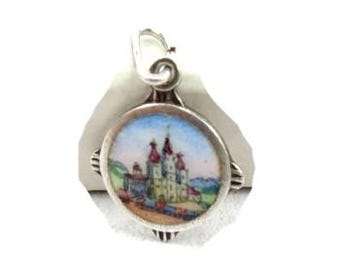 May include: A silver charm with a hand-painted enamel scene of a castle with red roofs and a church steeple. The charm is round and has a detailed design.