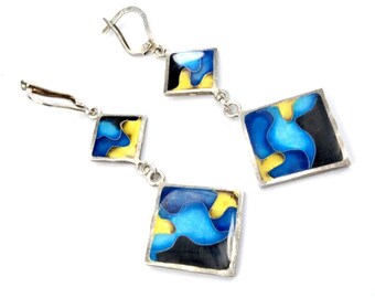 May include: A pair of silver dangle earrings with three square-shaped pendants. Each pendant features a blue, yellow, and black abstract design.