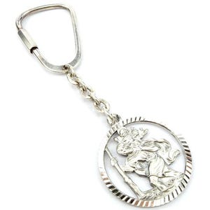 Op de afbeelding: Een zilverkleurige sleutelhanger met een gedetailleerde afbeelding van Sint-Christoffel die het Christuskind draagt. De ronde hanger heeft een getextureerde rand en is bevestigd aan een ketting en een karabijnhaak.