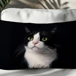 May include: Black rectangular pillow featuring a close-up of a black and white cat with green eyes. The cat has a white chest and face, with black markings around the eyes and ears. The pillow is on a white surface.