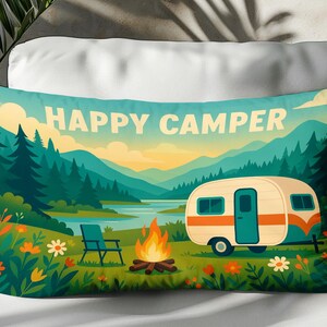 Può includere: Cuscino rettangolare con una scena di campeggio. Il cuscino presenta un camper, un falò e un paesaggio montano con la scritta "HAPPY CAMPER". La tavolozza dei colori include sfumature di verde, blu, arancione e bianco.