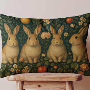 May include: Rectangular decorative pillow featuring four tan rabbits against a dark green background with yellow flowers and red apples. The rabbits are centered and facing forward. The pillow is placed on a wooden stool.
