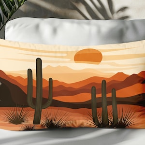 May include: Rectangular decorative pillow featuring a desert landscape scene. The design includes a setting sun, mountains, and cacti in shades of orange, brown, and tan. The pillow is placed on a white surface.