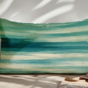 May include: Rectangular cushion featuring a horizontal striped design in teal, turquoise, and cream. The cushion is placed on a neutral surface, with shadows cast across it.