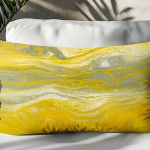 May include: A rectangular decorative pillow with a vibrant abstract design. The pillow features a flowing pattern of yellow, gray, and white hues, creating a marbled effect. The pillow is placed on a white surface.