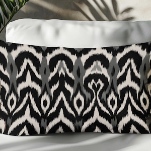 May include: Rectangular cushion with a black, grey, and off-white ikat pattern. The cushion is on a white surface, with a wicker chair and greenery in the background. The cushion's design features a repeating, stylized zigzag motif.