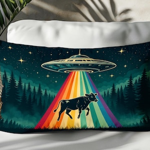 May include: A rectangular pillow with a whimsical design. The image features a black cow being beamed up by a UFO, with a rainbow beam of light. The background shows a forest under a starry night sky. The pillow is placed on a white surface.
