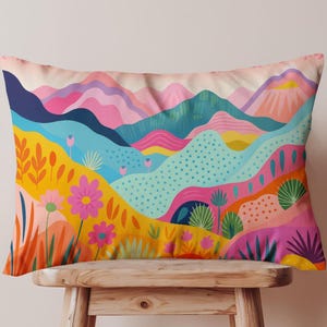 May include: Rectangular decorative pillow with a colorful landscape design. The pillow features a vibrant illustration of rolling hills, flowers, and mountains in shades of pink, blue, yellow, and orange. The pillow is resting on a wooden stool.
