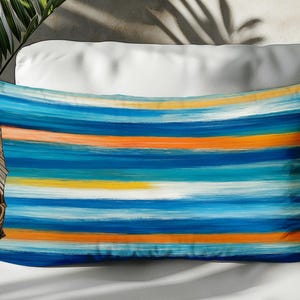 May include: Rectangular throw pillow with a vibrant abstract design. The pillow features horizontal brushstrokes in shades of blue, orange, yellow, and white. The pillow is placed on a white surface, with a wicker chair and plant in the background.