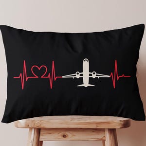 May include: Black rectangular pillow featuring a red heartbeat line with a heart shape and a white airplane graphic. The pillow is resting on a wooden stool. The design is simple and graphic.