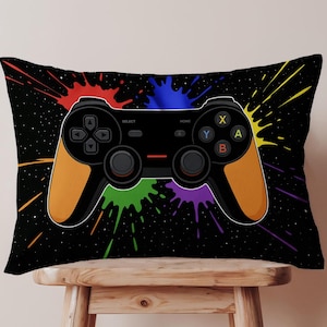 Könnte beinhalten: Ein schwarzes rechteckiges Kissen mit einem Videospiel-Controller-Design. Der Controller ist schwarz mit orangefarbenen Griffen und bunten Knöpfen. Der Hintergrund ist schwarz mit bunten Farbspritzern.
