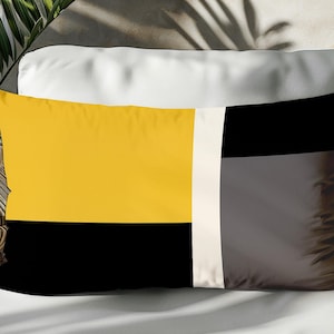 Può includere: Cuscino decorativo rettangolare con un design geometrico. Il cuscino presenta un motivo a blocchi di colore giallo, nero e grigio con una striscia bianca verticale. Il cuscino è appoggiato su una superficie bianca.