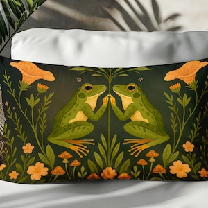 Op de afbeelding: Rechthoekig decoratief kussen met een grillig ontwerp. Twee groene kikkers staan tegenover elkaar, omringd door oranje bloemen, paddenstoelen en bladeren tegen een donkergroene achtergrond. Het kussen ligt op een wit oppervlak.