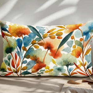 Può includere: Cuscino rettangolare decorativo con un motivo floreale ad acquerello. Il design presenta fiori e foglie arancioni, gialli, blu e verdi su sfondo bianco. Il cuscino è posto su una superficie dai toni neutri.