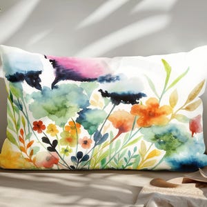 Può includere: Cuscino rettangolare decorativo con un design floreale ad acquerello. Il cuscino presenta una vibrante gamma di fiori nei toni dell'arancione, giallo, blu e verde su uno sfondo bianco. È un oggetto decorativo per un letto o un divano.