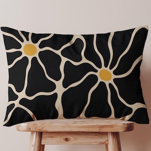 Op de afbeelding: Een rechthoekig decoratief kussen met een zwarte achtergrond en een herhalend bloemenpatroon. De bloemen hebben gele centra en gebroken witte bloemblaadjes. Het kussen ligt op een kleine houten kruk.