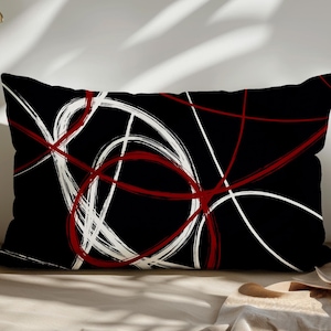 May include: Rectangular black pillow with a modern abstract design. The design features overlapping lines in white and red, creating a dynamic and artistic pattern. The pillow is likely for home decor.