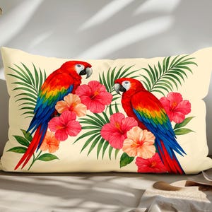 Op de afbeelding: Rechthoekig crèmekleurig kussen met twee rode ara's met blauwe en gele vleugels, omringd door roze hibiscusbloemen en groene palmbladeren. Een decoratief item voor in huis met een tropisch thema.