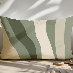 Op de afbeelding: Rechthoekig decoratief kussen met een golvend gestreept patroon in groen- en beigetinten. Het kussen ligt op een neutrale ondergrond. Een houten lepel en een opgevouwen doek liggen op de voorgrond. Het ontwerp voegt een moderne touch toe.