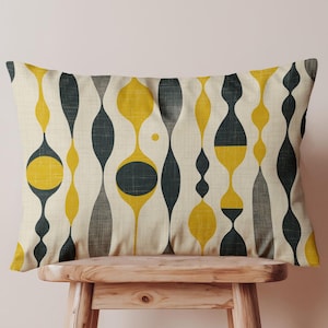 May include: Rectangular decorative pillow with a mid-century modern design. The pillow features a repeating pattern of yellow, gray, and dark teal shapes on a cream-colored background. The pillow is displayed on a small wooden stool.