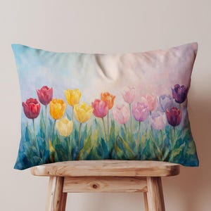Op de afbeelding: Rechthoekig decoratief kussen met een aquarel schilderij van kleurrijke tulpen. De bloemen zijn in tinten rood, geel, oranje, roze en paars, tegen een zachte blauwe en roze achtergrond. Het kussen ligt op een houten kruk.
