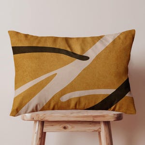 May include: Mustard-colored rectangular pillow with an abstract design. The design features black, white, and beige curved lines across the pillow's surface. The pillow is resting on a small, light-colored wooden stool.