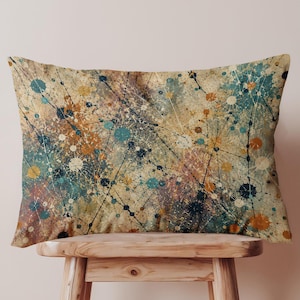 May include: A rectangular throw pillow featuring an abstract pattern of splattered paint in shades of teal, mustard yellow, and dark brown against a beige background, with thin black lines connecting various circular paint spots across the surface.
