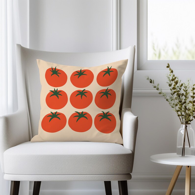 May include: A decorative throw pillow featuring a repeating pattern of nine red tomatoes with green stems on a cream-colored background. The pillow is square and rests on a white armchair, suggesting a home decor theme.