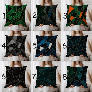May include: A selection of square throw cushions with geometric patterns. The cushions display various colour schemes, including green, blue, orange, and grey, with gold lines forming the geometric designs. The cushions appear to be made of a soft material and are held up by a person.