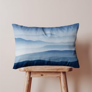 Op de afbeelding: Een rechthoekig kussen met een blauw bergketenontwerp. Het kussen heeft lagen blauwtinten, waardoor een gradiënteffect ontstaat. Het kussen ligt op een houten kruk.