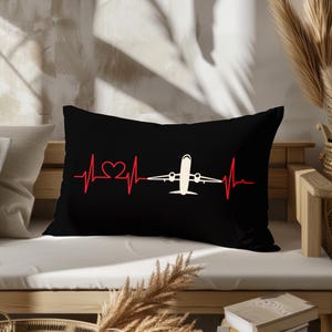 May include: Black rectangular pillow with a red heartbeat line design that includes a heart and a white airplane. The pillow is on a white surface and a wooden bench.