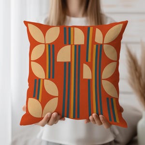 May include: Square orange decorative pillow with a geometric pattern. The design features beige petal shapes and vertical stripes in navy blue and yellow. The pillow is held by a person.