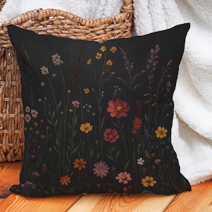 Può includere: Cuscino nero quadrato con un vivace motivo floreale. Il design presenta una varietà di fiori selvatici in tonalità rosse, gialle, rosa e viola, su uno sfondo scuro. Il cuscino sembra essere fatto di tessuto morbido.