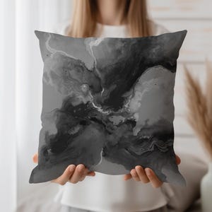 May include: Square cushion with a black and grey abstract design. The cushion features a marbled effect with swirling patterns in shades of grey and black. The cushion is held up by a person.