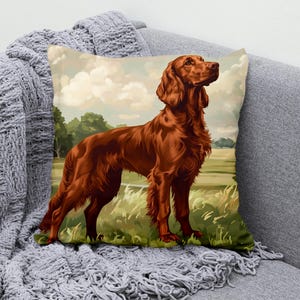 Peut inclure: Un coussin carré orné d'une illustration d'un Setter irlandais dans un champ. Le chien est représenté dans des tons de marron et de rouge, avec un arrière-plan d'herbe verte, d'arbres et d'un ciel nuageux. Le coussin est posé sur un canapé gris.