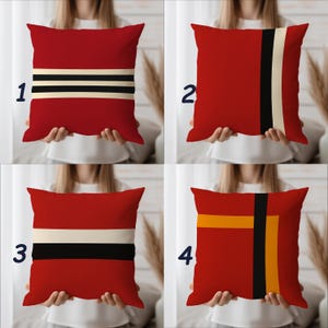 May include: Four red decorative throw pillows with geometric designs. Each pillow features a different pattern: stripes, vertical lines, and a cross design. The designs incorporate black, white, and orange accents. The pillows are square and appear to be made of a soft fabric.