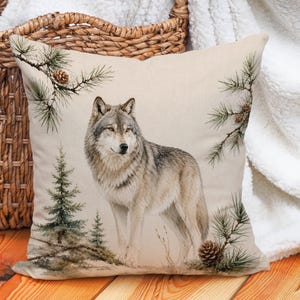May include: A square throw pillow featuring a detailed illustration of a gray and white wolf standing in a forest setting. The design includes green pine branches with brown pinecones and small evergreen trees against a light beige background.