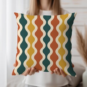 May include: Square decorative pillow with a retro wavy pattern in teal, mustard yellow, rust orange, and cream colors. The pillow is a square shape and appears to be made of a soft fabric. The design is a repeating vertical pattern.
