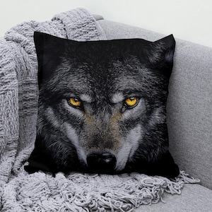 May include: A black and grey wolf cushion with bright yellow eyes. The wolf's face is close up and the background is black.