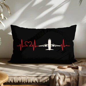 May include: Black rectangular pillow with a red heartbeat line design and a white airplane in the center. The heartbeat line includes a heart shape. The pillow is on a wooden surface.