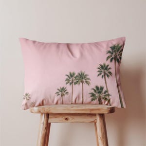 Op de afbeelding: Een rechthoekig roze kussen met een print van palmbomen. Het kussen heeft een zachte textuur en staat op een houten kruk. De palmbomen zijn groen met bruine stammen, tegen een roze achtergrond.