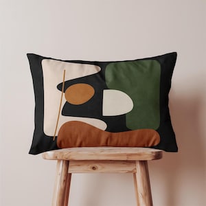 May include: Rectangular decorative pillow with an abstract design. The pillow features a black background with shapes in beige, brown, green, and white. The pillow is resting on a wooden stool.