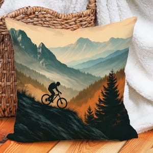 May include: Square cushion with a mountain biking scene. The design depicts a cyclist on a mountain trail with layered blue and green mountains under a warm, orange sky. The cushion is placed on a wooden surface, with a wicker basket in the background.