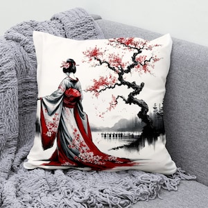 Könnte beinhalten: Ein weißes Kissen mit einer Schwarzweißillustration einer Frau in einem rot-weißen Kimono, die vor einem Schwarzweiß-Kirschblütenbaum steht. Der Hintergrund ist eine Schwarzweißlandschaft mit einem See und einer Brücke.