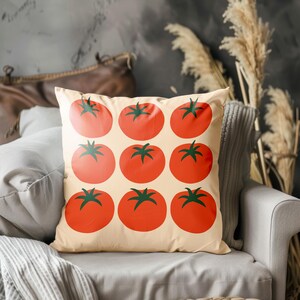 May include: Square throw pillow with a cream background featuring a pattern of nine large, red tomatoes with green stems. The pillow is on a gray chair with a gray blanket. Decorative items are in the background.