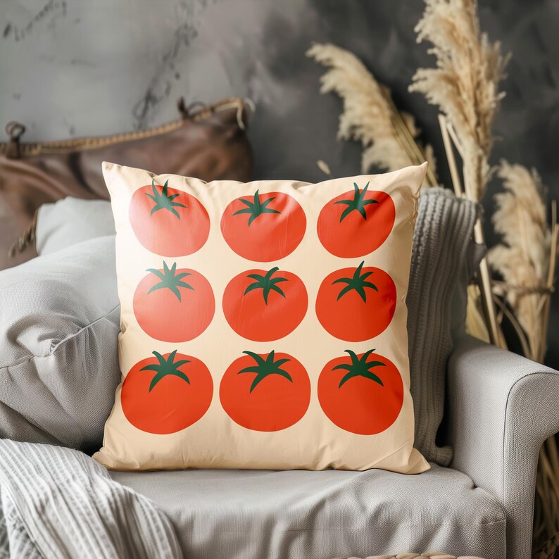 May include: Square throw pillow with a cream background featuring a pattern of nine large, red tomatoes with green stems. The pillow is on a gray chair with a gray blanket. Decorative items are in the background.